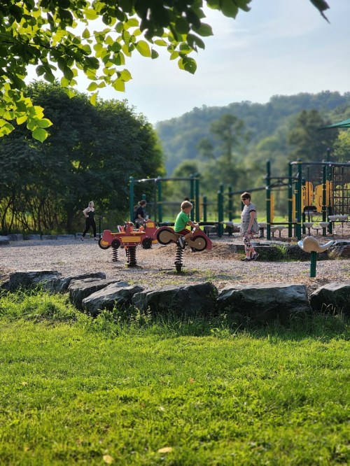 Children playing on spring riders at a playground, surrounded by greenery and hills in the background.