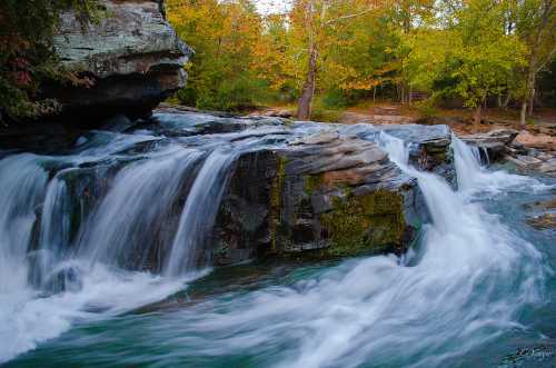 A serene waterfall cascades over rocky ledges, surrounded by vibrant autumn foliage and a tranquil forest setting.