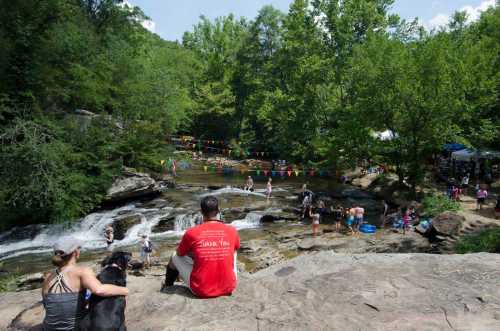 A scenic river with people swimming and playing, surrounded by trees and colorful flags, with a couple sitting on a rock.