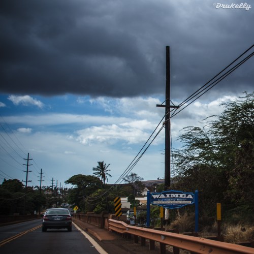 A scenic road in Waimea, with cloudy skies and power lines, surrounded by trees and distant hills.