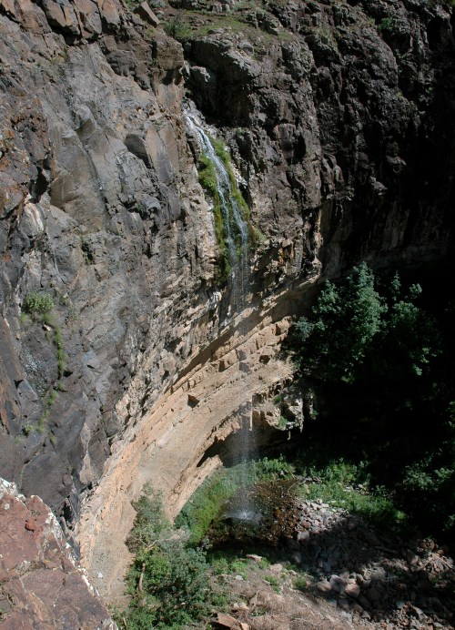 A waterfall cascading down a rocky cliff into a lush green area below, surrounded by steep rock formations.