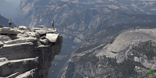 Two people stand on a rocky cliff overlooking a vast mountainous landscape.