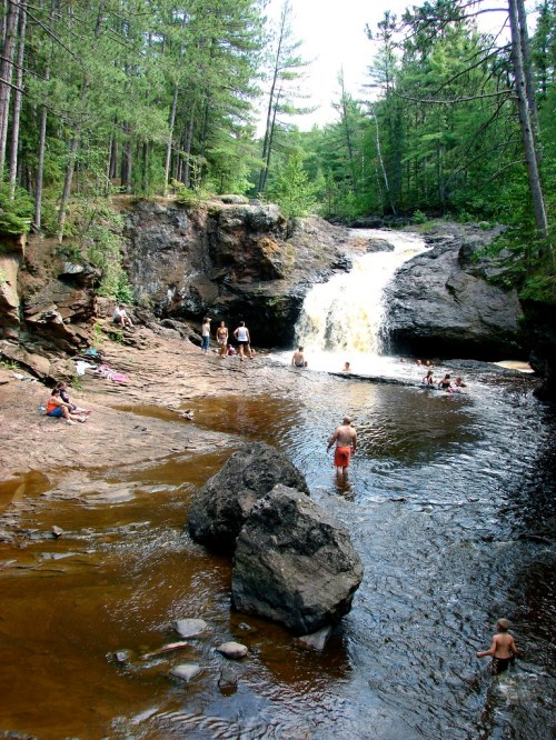 A scenic waterfall surrounded by lush trees, with people swimming and relaxing in the water below.