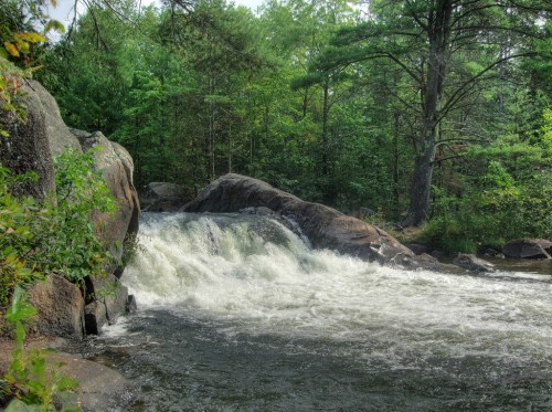A serene waterfall cascades over rocks, surrounded by lush green trees and a tranquil river.