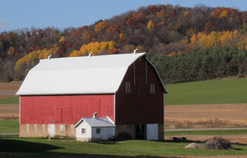 A red barn with a white roof stands in a green field, surrounded by autumn-colored trees on a hillside.