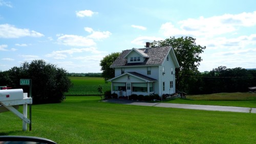 A white two-story house with a porch, surrounded by green grass and fields under a blue sky with fluffy clouds.