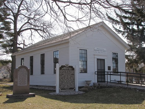A small white historic building with large windows, surrounded by trees and a grassy area, featuring informational plaques outside.