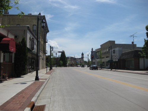A quiet street scene with shops and buildings under a clear blue sky, featuring traffic lights and a few parked cars.