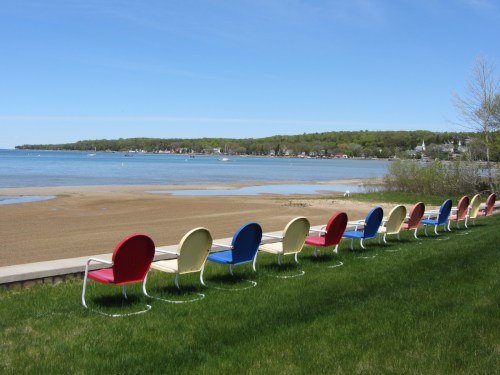 Colorful chairs line a grassy area by a calm lake, with a sandy beach and green hills in the background.