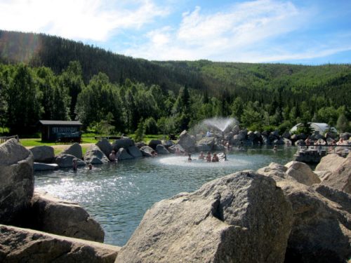 A natural hot spring surrounded by large rocks and lush greenery, with people enjoying the water under a blue sky.