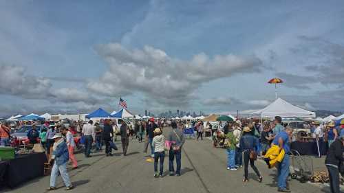 A bustling outdoor market with tents, people walking, and a city skyline in the background under a cloudy sky.