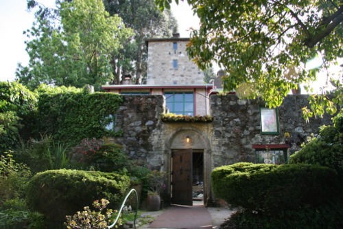 A stone building surrounded by lush greenery and trees, featuring an arched entrance and colorful windows.