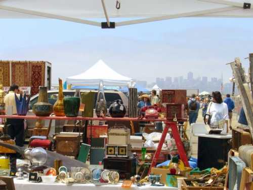 A bustling outdoor flea market with various vintage items for sale and a city skyline in the background.