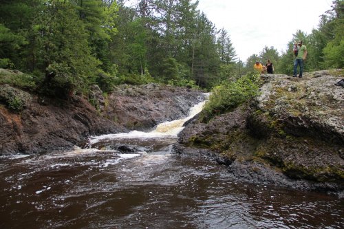 A rocky riverbank with a small waterfall surrounded by lush greenery and two people standing nearby.