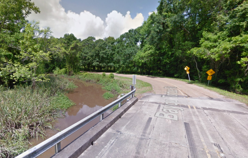 A rural road curves near a small creek, surrounded by lush greenery and trees under a partly cloudy sky.