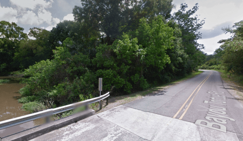 A winding road alongside a river, surrounded by lush greenery and trees under a cloudy sky.