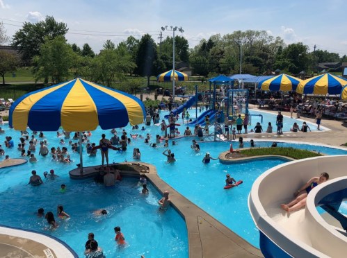 A busy water park with colorful umbrellas, slides, and many people enjoying the pool on a sunny day.