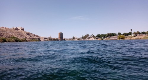 A view of a river with buildings and palm trees along the shore under a clear blue sky.