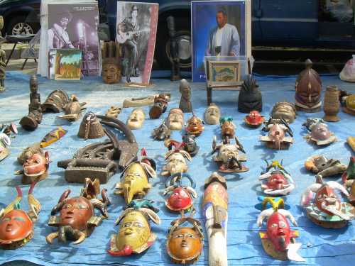 A display of colorful masks and sculptures on a blue tarp, with framed photos in the background.