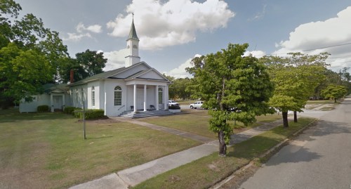 A white church with a steeple, surrounded by trees and a grassy area, located on a quiet street.