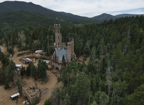 Aerial view of a stone castle surrounded by trees and mountains, with vehicles and people nearby.