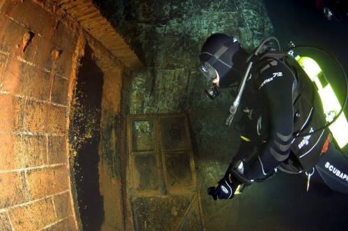 A diver explores a submerged, rusted structure with brick walls and a window frame, surrounded by dark water.