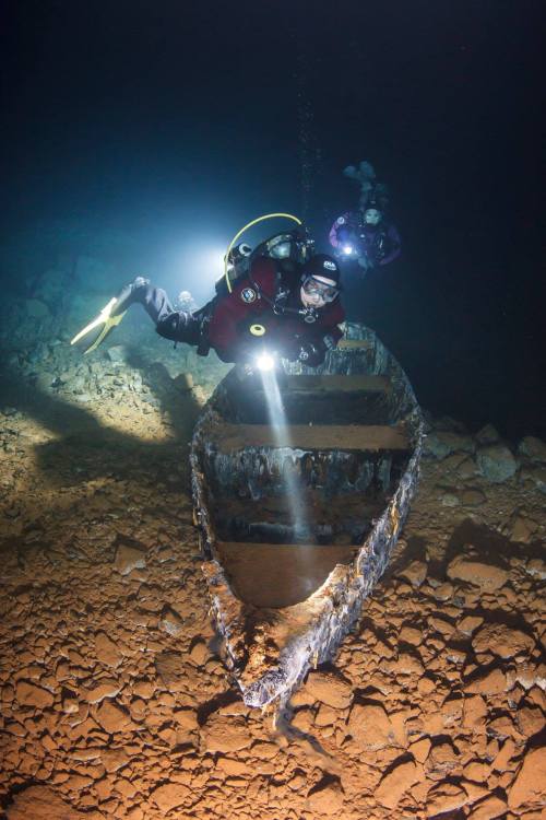 Divers explore a sunken shipwreck underwater, illuminated by their lights in a dark, rocky environment.
