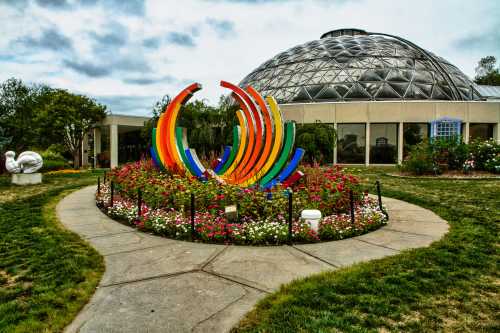 Colorful sculpture surrounded by flowers in a garden, with a large glass dome building in the background.