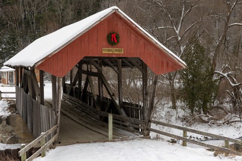 A snow-covered covered bridge with a red roof and a wreath, surrounded by trees in winter.