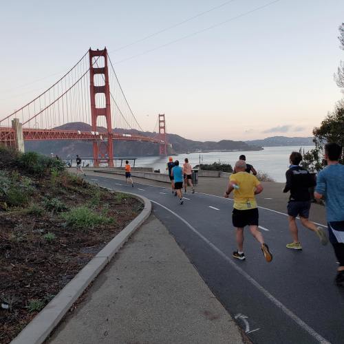 Runners on a path near the Golden Gate Bridge at sunset, with hills and water in the background.