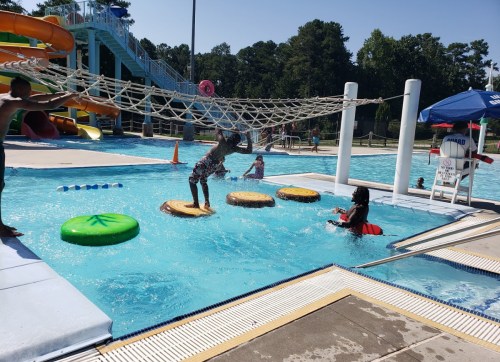Children playing in a pool with floating obstacles and a water slide in the background on a sunny day.
