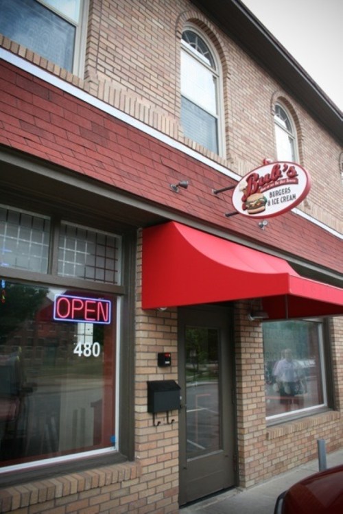A brick building with a red awning and a sign reading "Benny's Burgers & Ice Cream," featuring an "Open" neon sign.