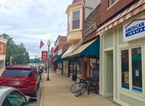 A quaint street scene featuring shops, parked cars, and a bicycle near a bench, with an American flag in the background.