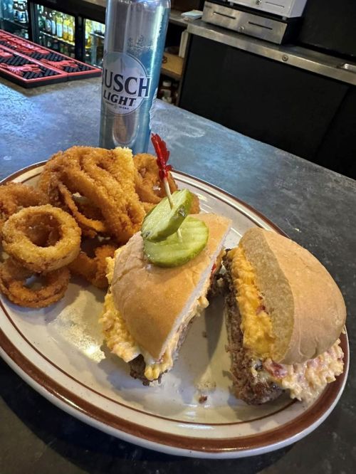 A plate with a sandwich filled with creamy toppings, a pickle, and a cherry, alongside onion rings and a can of beer.