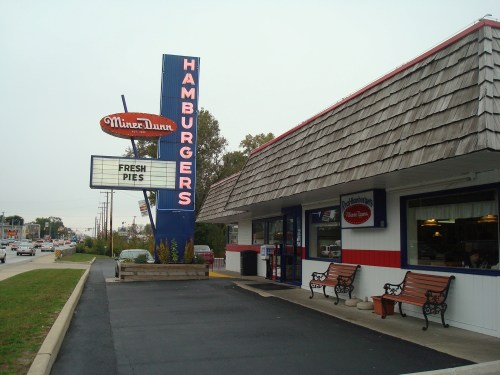 A retro-style hamburger restaurant with a sign advertising "Fresh Pies" and a shingled roof, surrounded by benches.