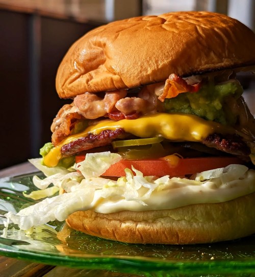 A close-up of a delicious burger with layers of cheese, bacon, lettuce, tomato, and guacamole on a toasted bun.