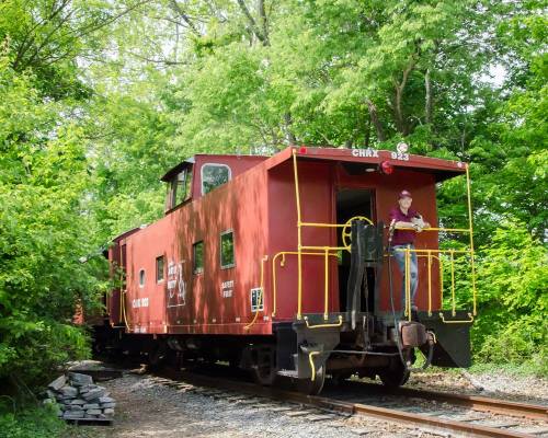 A red caboose parked on a track, surrounded by lush green trees, with a person standing on the platform.