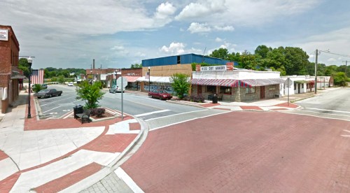 A small town street scene with shops, trees, and a blue sky, featuring a red brick road and parked cars.