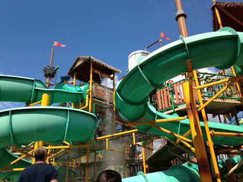 A colorful water park with green slides and a wooden play structure under a blue sky. People are walking towards it.