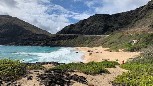 A scenic beach with golden sand, turquoise water, and lush green hills under a partly cloudy sky. People stroll along the shore.