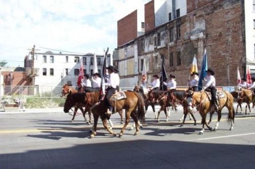 A group of horse riders in traditional attire parades down a street, carrying flags against a backdrop of old buildings.