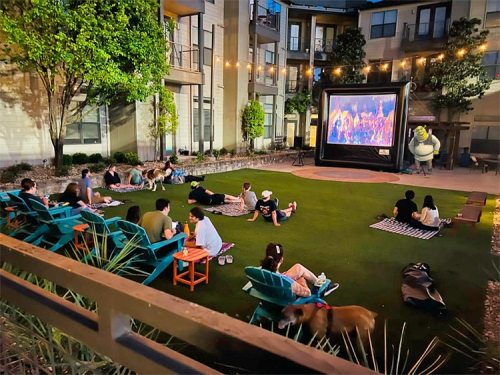 Outdoor movie night in an apartment courtyard, with people lounging on grass and chairs, watching a large screen.