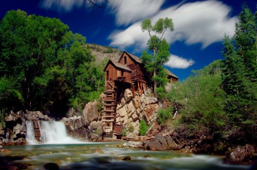 A historic wooden mill beside a waterfall, surrounded by lush green trees and a bright blue sky with clouds.
