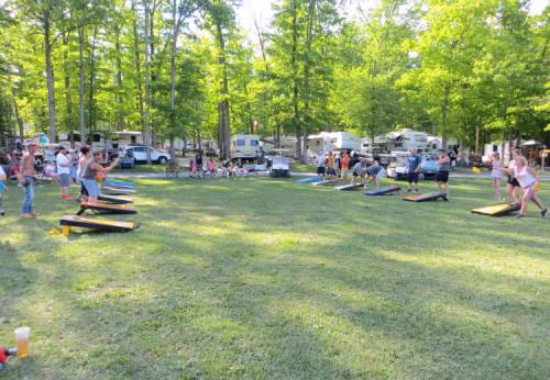 A lively outdoor scene with people playing cornhole in a grassy area surrounded by trees and campers.