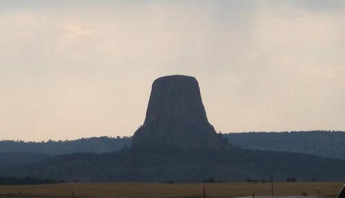 A large, flat-topped rock formation rises above a landscape, surrounded by trees and a cloudy sky.
