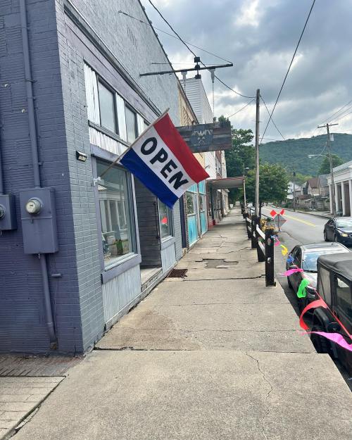 A colorful "OPEN" sign hangs outside a shop on a quiet street, with mountains in the background and decorative streamers.