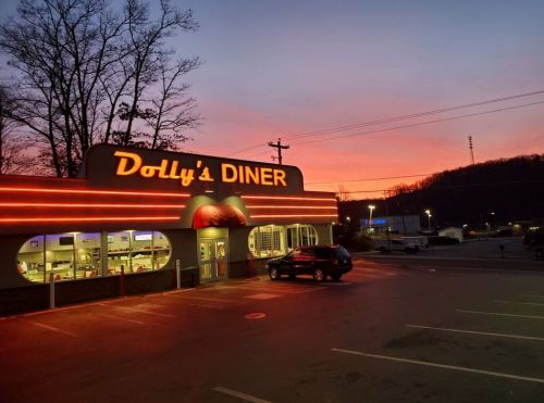 A diner with neon lights at sunset, surrounded by trees and a parking lot. The sky is painted in vibrant colors.