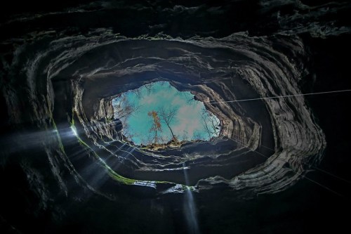 View from inside a cave looking up at a circular opening, with sunlight streaming through and trees visible above.
