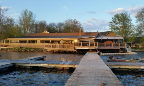 A lakeside restaurant with a wooden deck and dock, surrounded by trees and water under a blue sky.