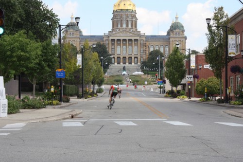 A cyclist rides down a street with a view of a grand building and dome in the background, surrounded by trees.